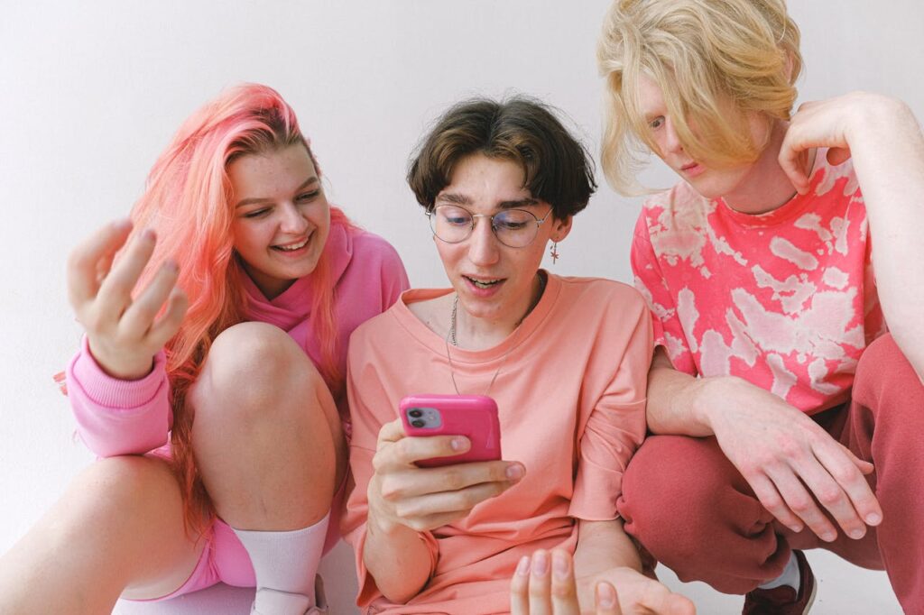 Three happy teenagers sitting together indoors, smiling and looking at a smartphone, enjoying social media or online content.