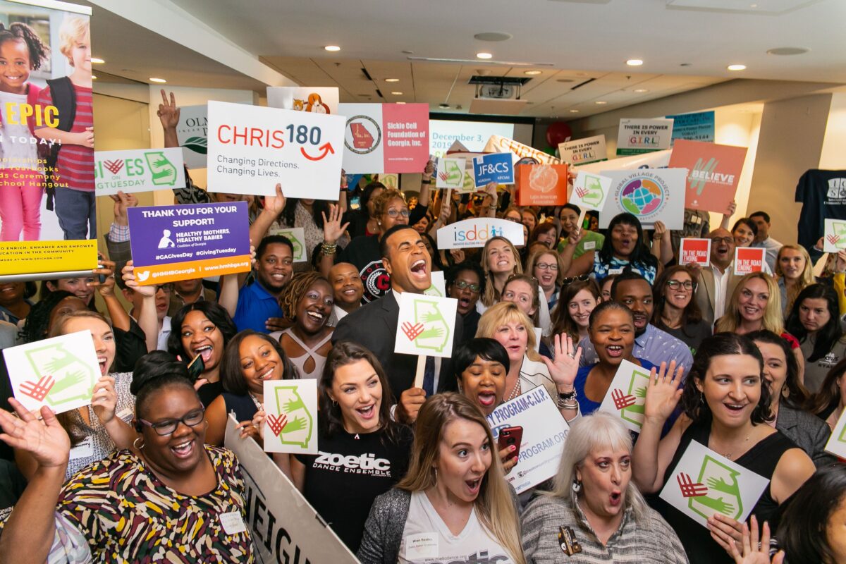 A lively gathering of people indoors celebrating a charity event with various signs and expressions of joy.